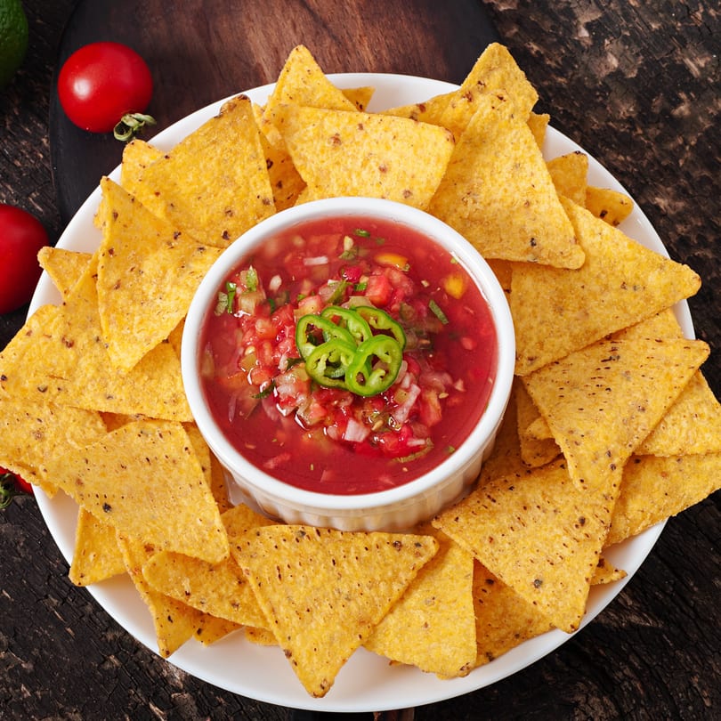 A white plate of golden tortilla chips surrounds a bowl of fresh red salsa with green peppers.