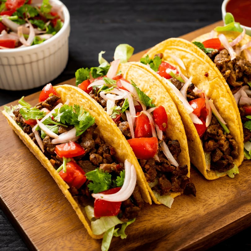 Four Metztli Mexican Taqueria tacos with meat, lettuce, tomatoes, onions, and cilantro on a wooden board.