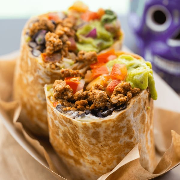 Two Metztli Mexican Taqueria burritos, filled with meat, beans, guacamole, and salsa, in a paper tray.