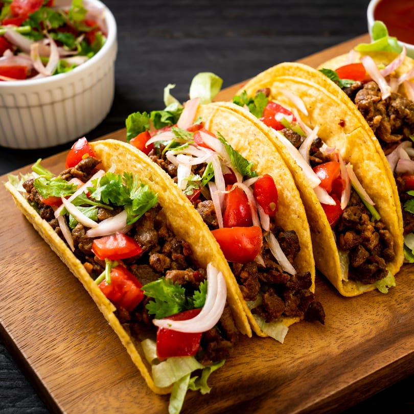 Three Metztli Mexican Taqueria tacos with meat, lettuce, tomatoes, onions, and cilantro on a wooden board.