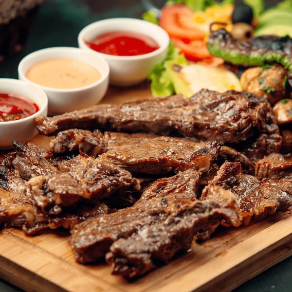 Grilled steak slices on a wooden board with three dipping sauces and a side salad.