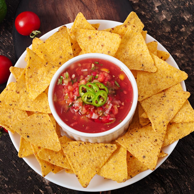 A white plate of golden tortilla chips surrounds a bowl of fresh red salsa with green jalapeños.