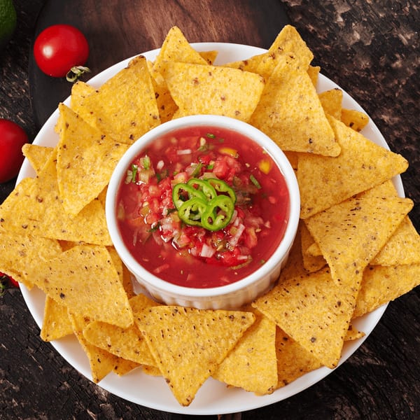 A white plate of golden tortilla chips surrounds a bowl of fresh red salsa, garnished with green peppers.