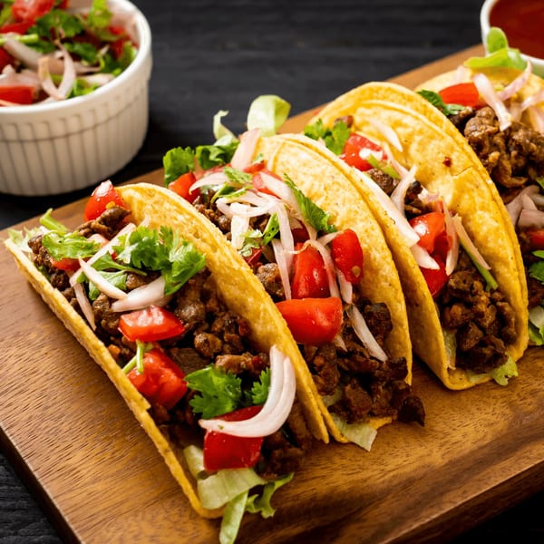 Four Metztli Mexican Taqueria tacos with meat, lettuce, tomatoes, onions, and cilantro on a wooden board.