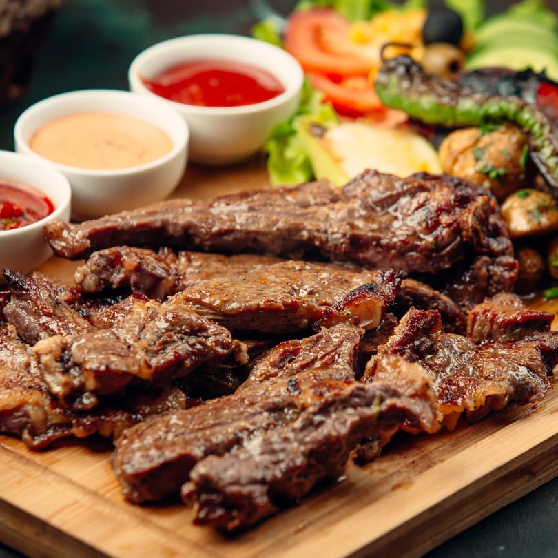 Grilled steak strips on a wooden board with three dipping sauces and a side salad.